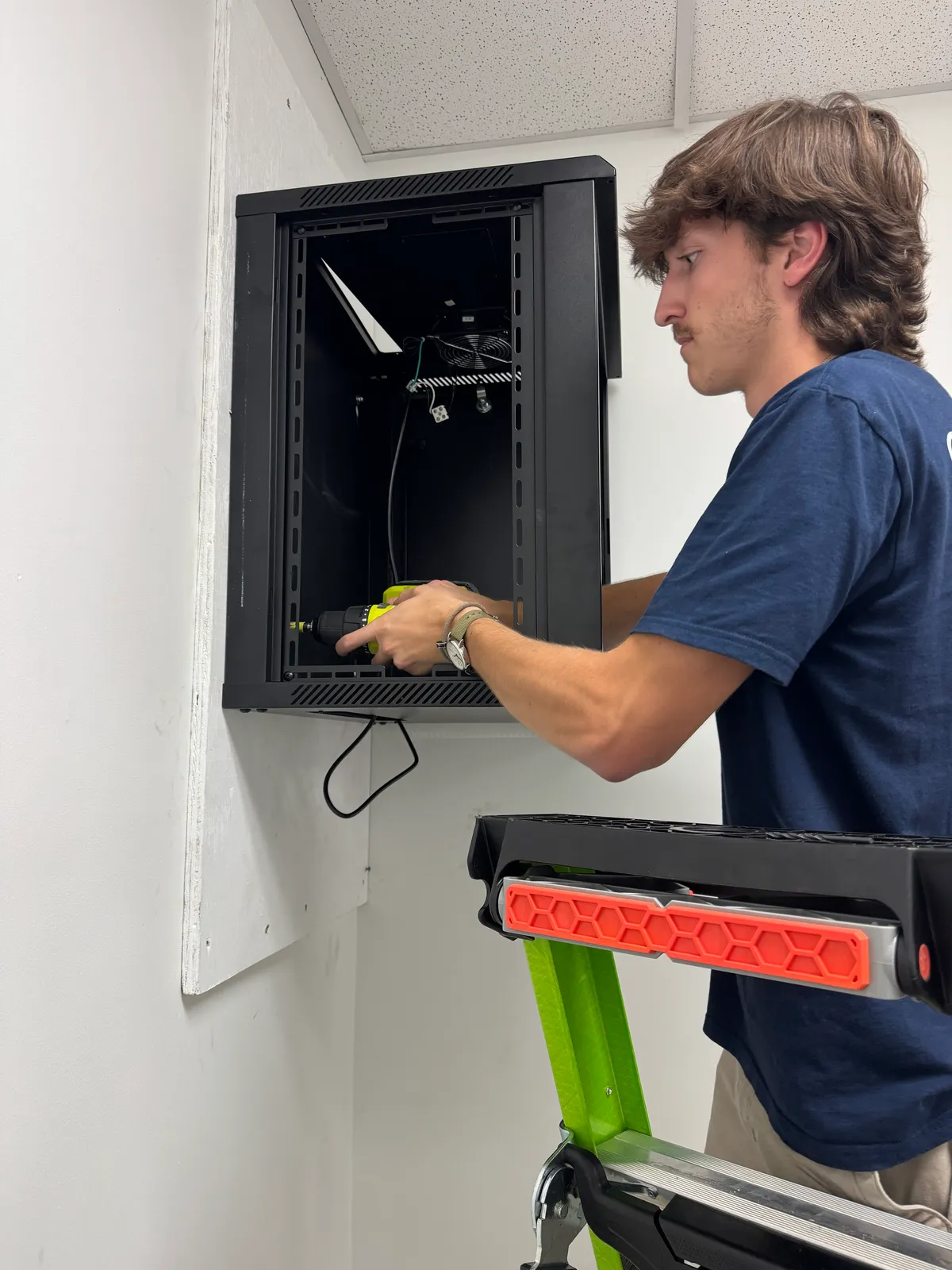 A Cyber Shift technician on a ladder installing a wall-mounted network cabinet A Cyber Shift technician on a ladder installing a wall-mounted network cabinet