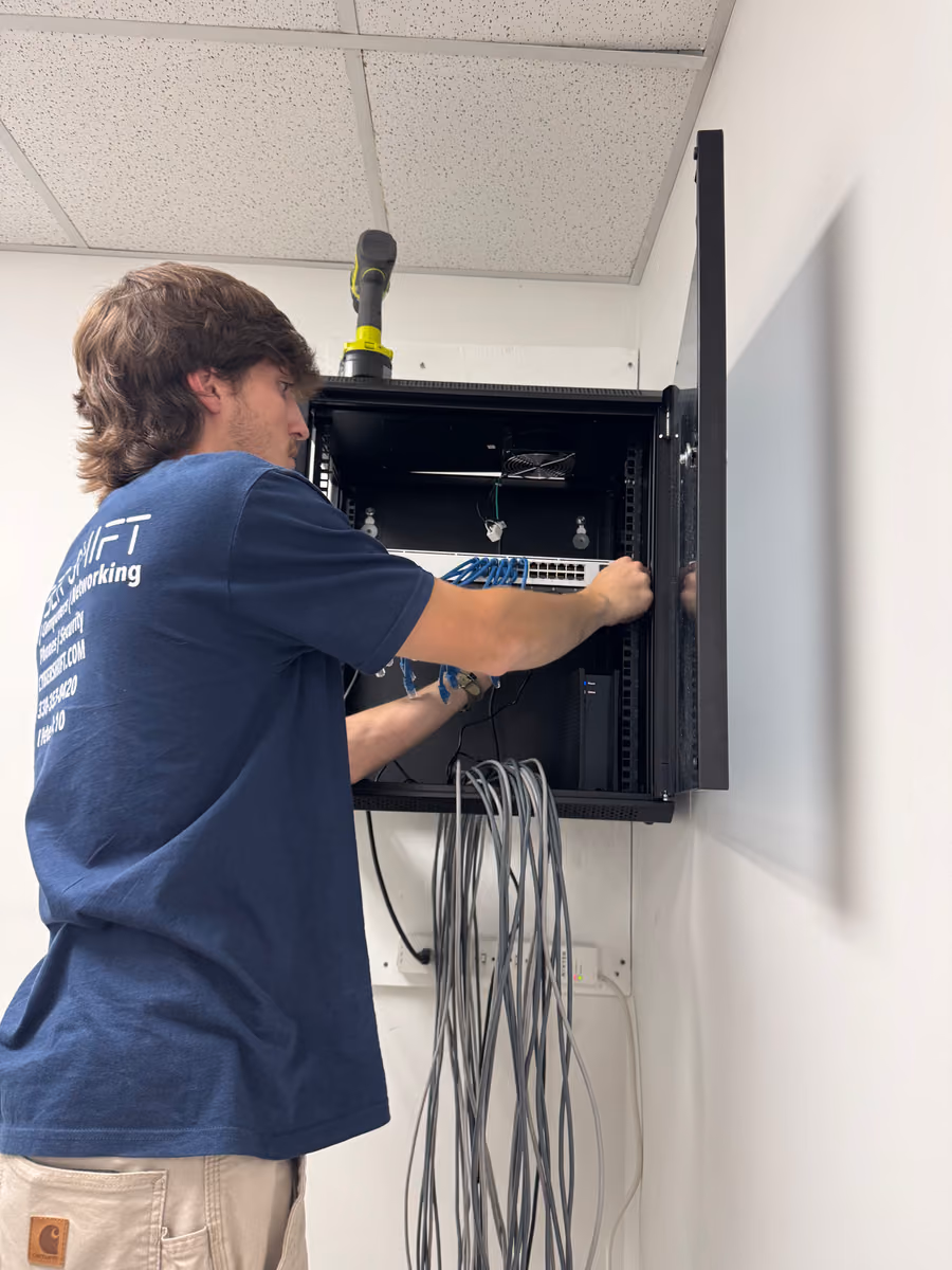 A Cyber Shift technician connecting blue patch cables to a switch in a wall-mounted network cabinet